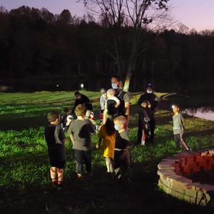 Cubs and Adults at a Campfire in a field by a lake in front of woods