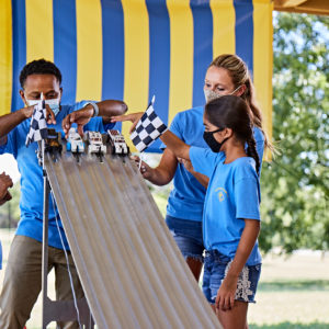 and Girl Cubs placing cars on Pinewood track in front of yellow and blue vertical striped curtain with tree in background.